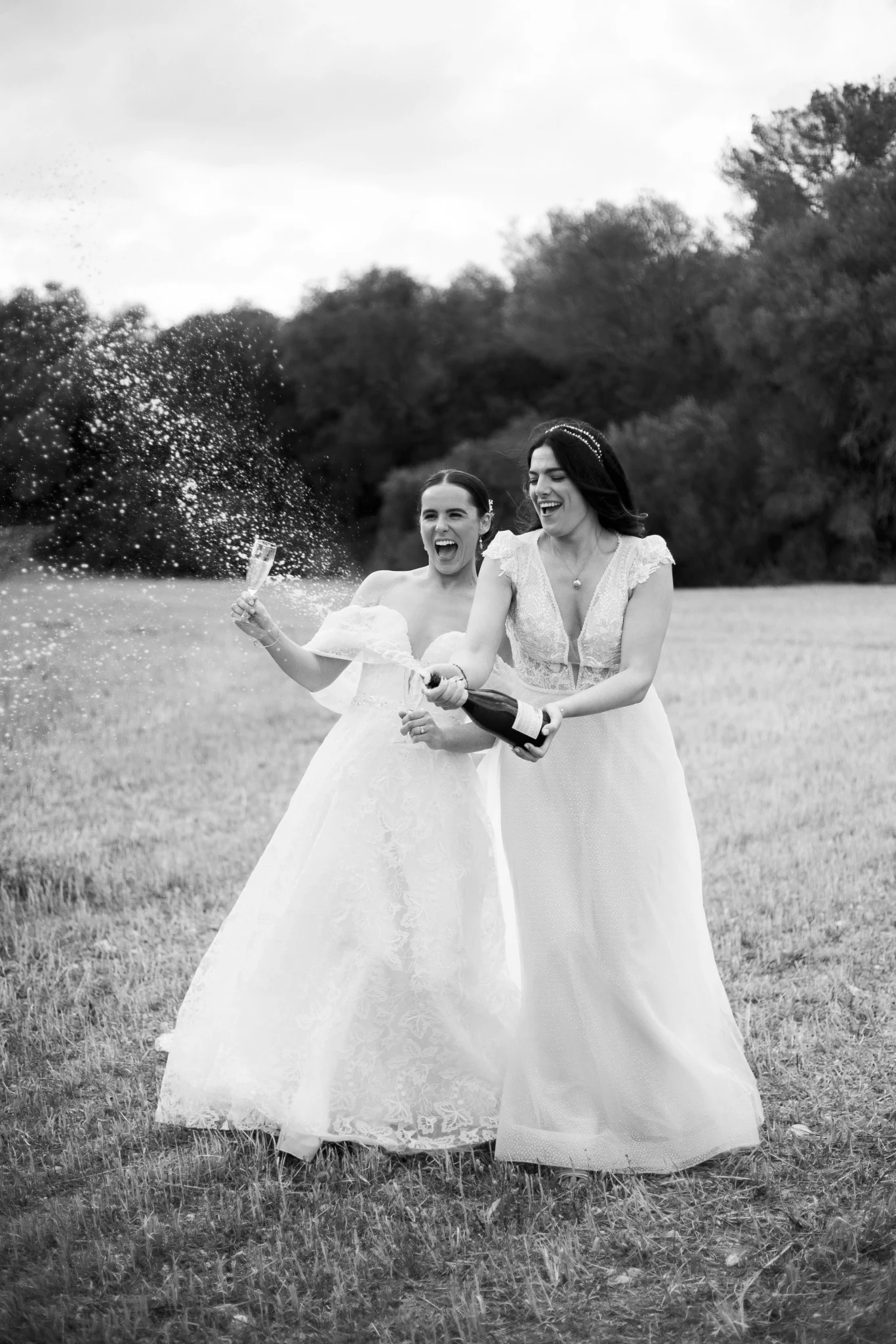 Two brides popping champagne in a field after their wedding – black and white photo by Inma del Valle