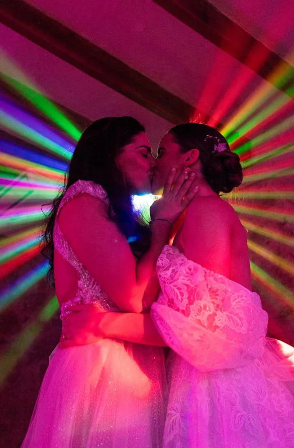 Two brides kissing under rainbow lights during their wedding party – photo by Inma del Valle
