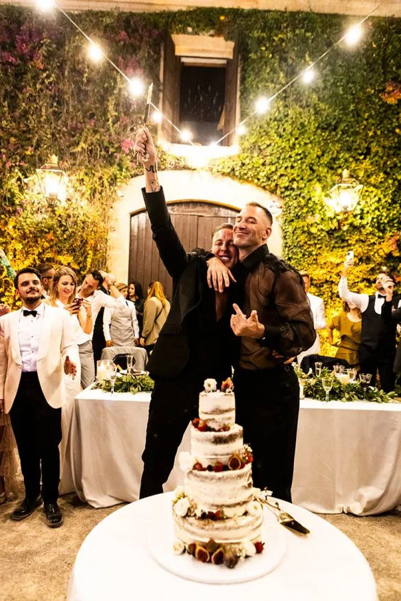 Pareja gay brindando junto a su tarta nupcial durante su boda de lujo en Mallorca, rodeados de invitados, fotografía de Inma del Valle.