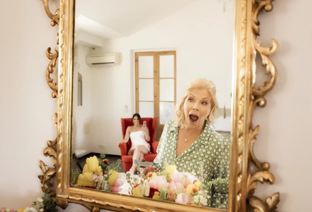 Bridesmaid reacts joyfully in front of mirror while bride relaxes in background before luxury wedding in Sitges.