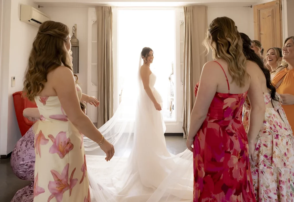 Bride illuminated by natural light, standing in her wedding dress, with bridesmaids admiring her in a luxury villa in Sitges.