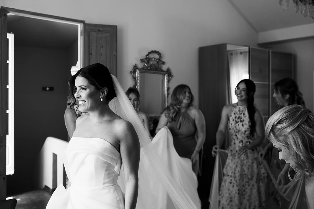 Bride smiling as she walks through the villa, followed by bridesmaids holding her dress in Sitges.