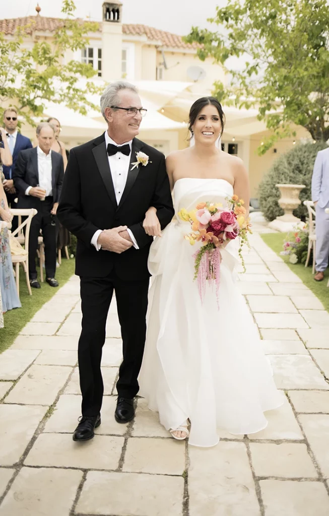 Bride holding a vibrant bouquet as she walks down the aisle with her father at a luxury wedding in Sitges.