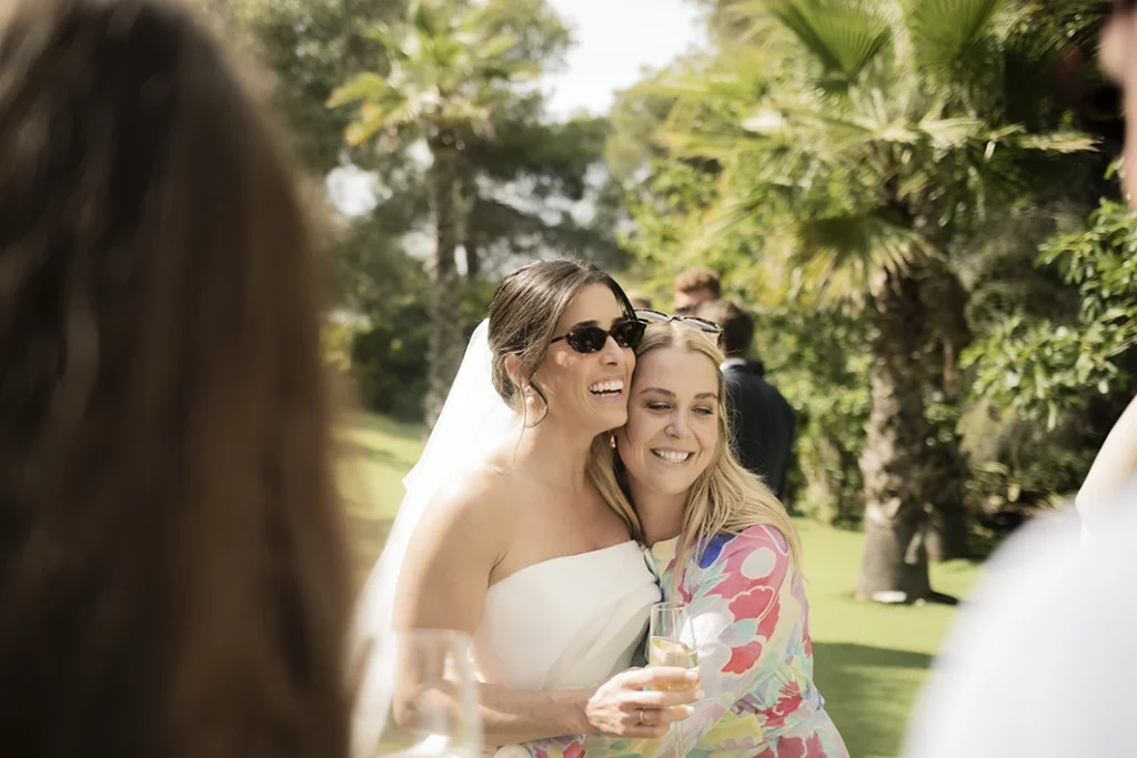 Bride wearing sunglasses hugging a smiling guest at a luxury wedding in Sitges.
