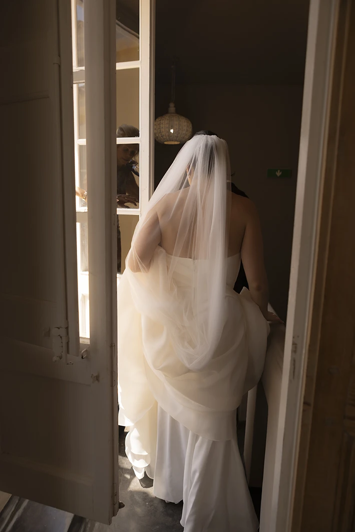 Bride walking toward the balcony in her wedding dress with veil, soft light entering the room in Sitges.