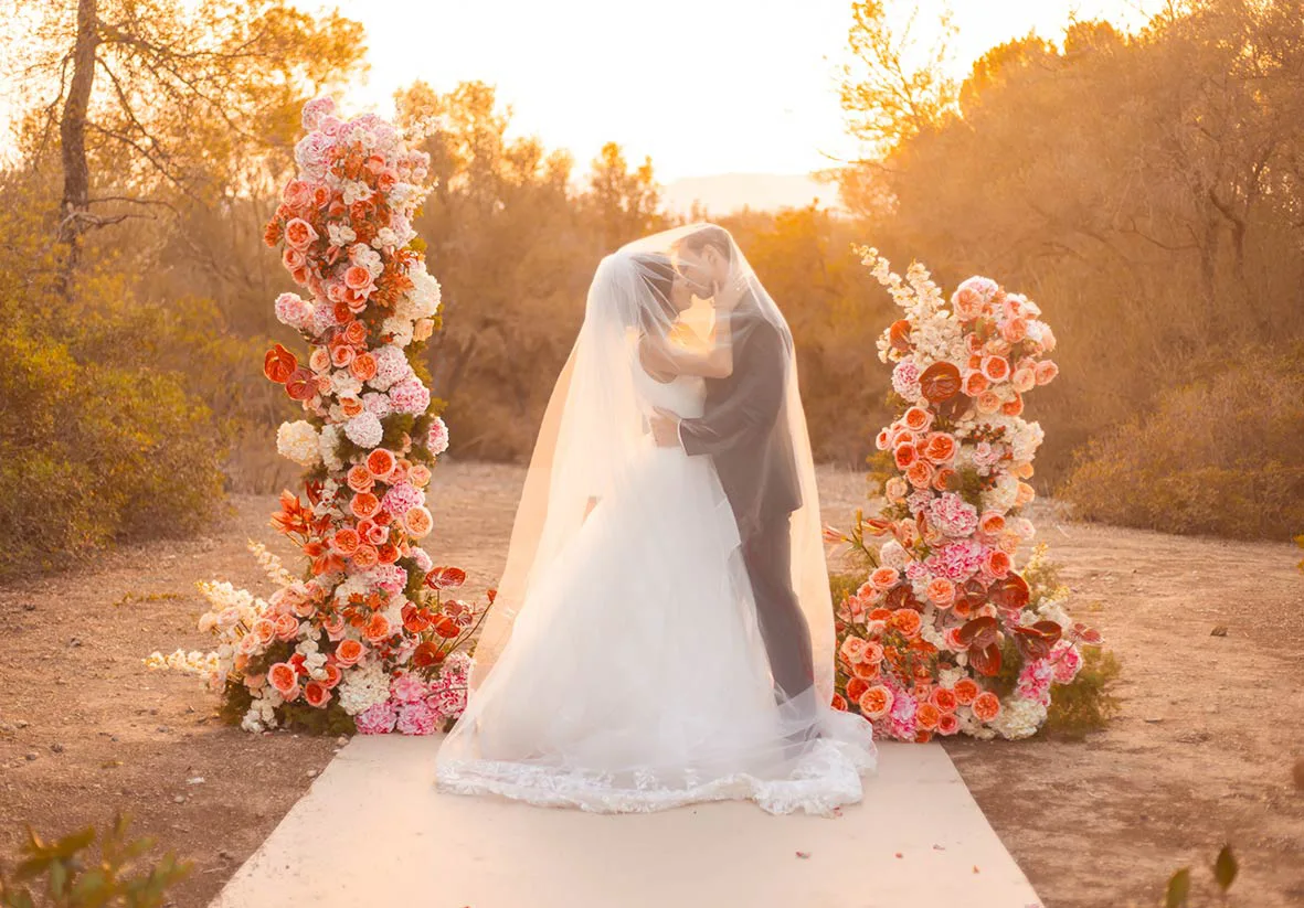 A breathtaking wedding moment captured at sunset, with the bride and groom sharing a kiss under a veil, surrounded by luxurious floral arrangements in soft pastel colors.