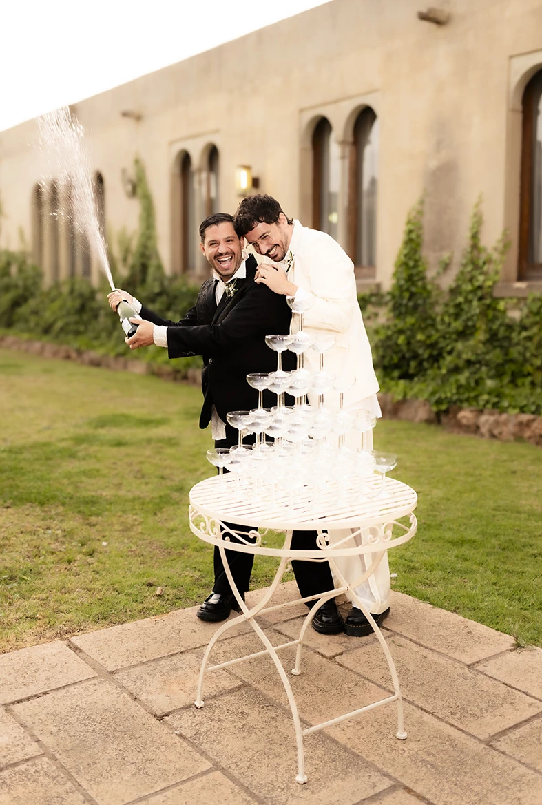 Jon y Santi descorchando una botella de champán junto a una torre de copas durante su boda en Finca Gordiola, Mallorca. Fotografía de Inma del Valle.