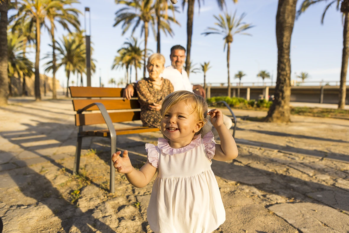 Niña pequeña sonriendo mientras corre en primer plano, con sus abuelos sentados en un banco al fondo rodeados de palmeras