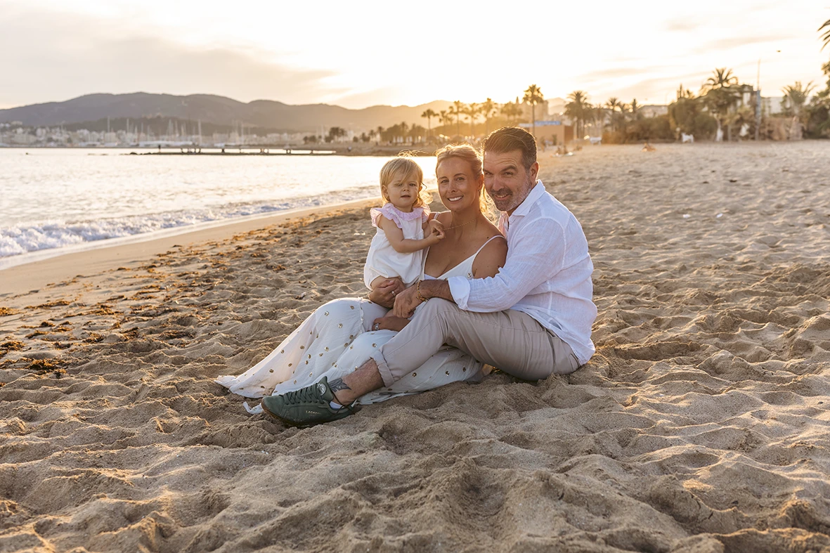 Familia sentada en la arena disfrutando del atardecer en la playa, con una niña pequeña en brazos