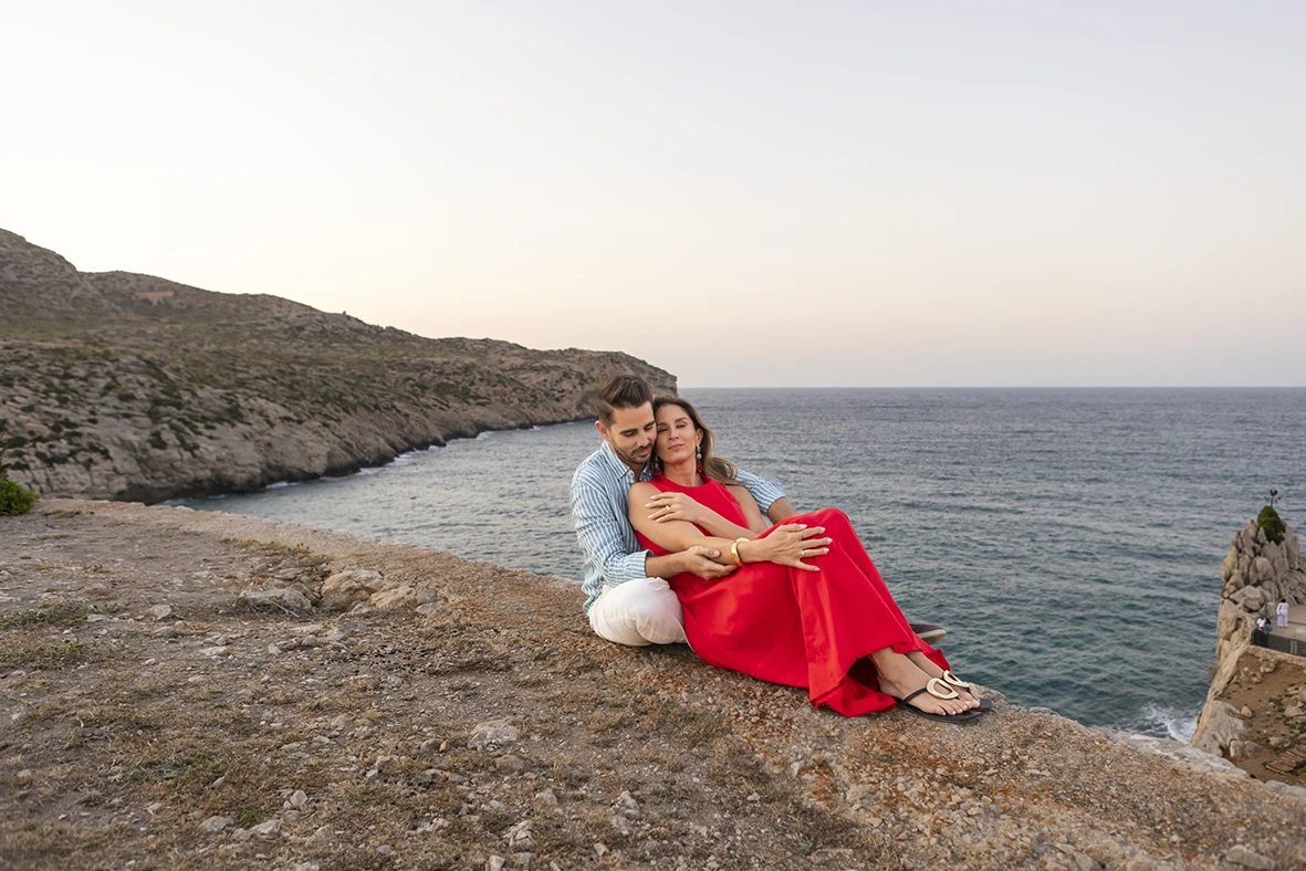 Couple sitting on cliffside at sunset after a surprise wedding proposal in Mallorca