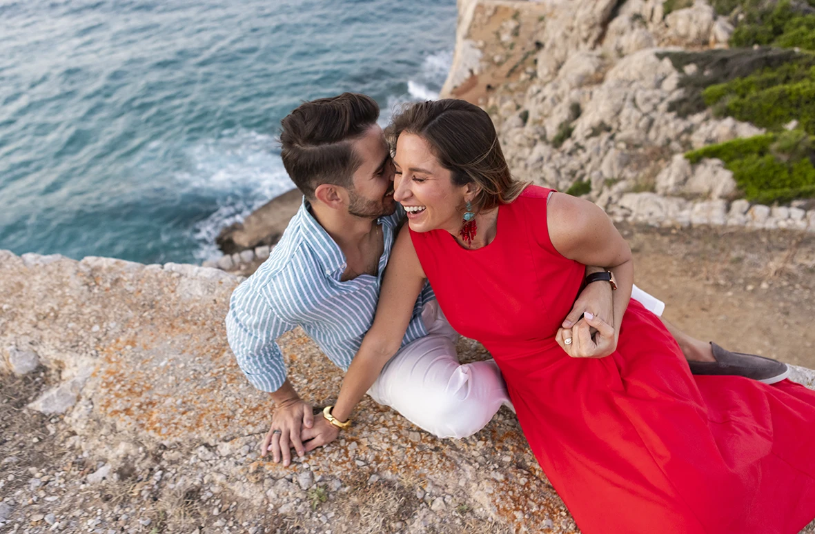 Playful couple sitting on a cliff after a surprise wedding proposal in Mallorca
