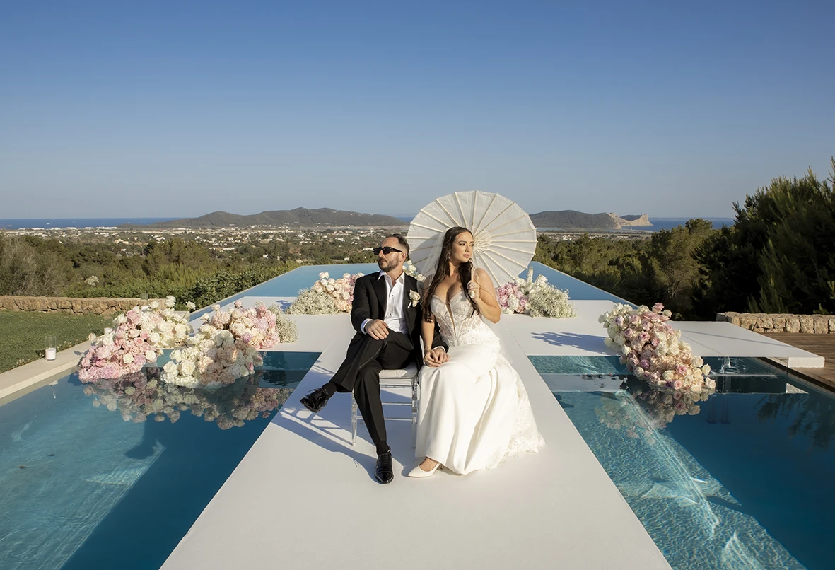 La pareja de recién casados sentados en la pasarela blanca sobre la piscina, ella sosteniendo una sombrilla con estilo, con Ibiza de fondo.