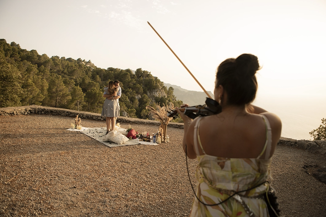Violinist playing while an emotional couple embraces after the proposal on a Mallorca cliff.