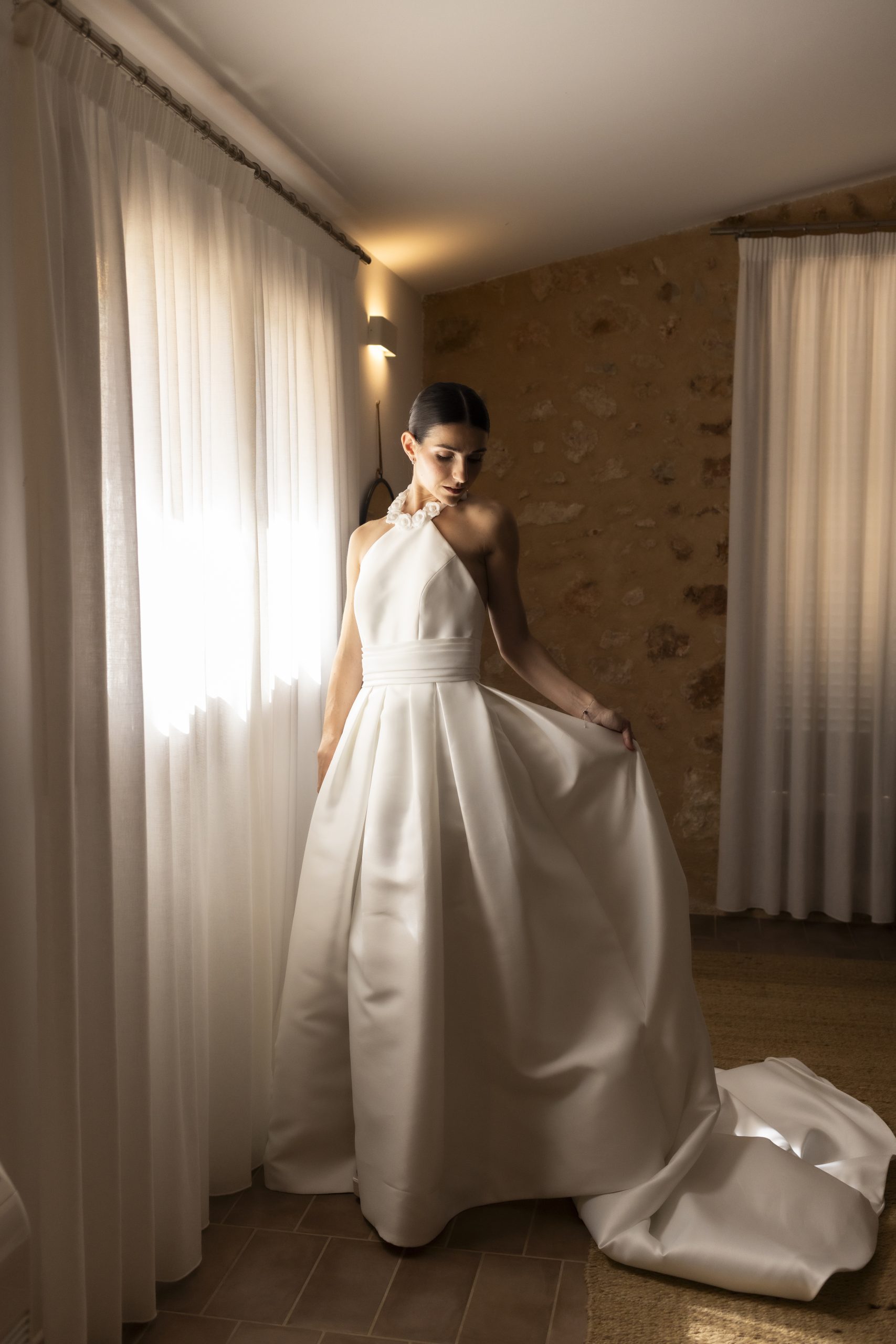 Bride in an elegant white wedding dress standing by the window with soft natural light in Mallorca.