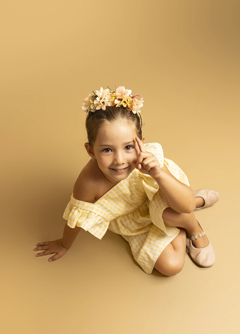 Niña con vestido amarillo y corona de flores señalando. Fotógrafo de niños para agencia en Mallorca.