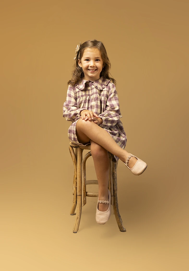 Niña con vestido de cuadros sentada en taburete. Fotógrafo de niños para agencia en Mallorca.
