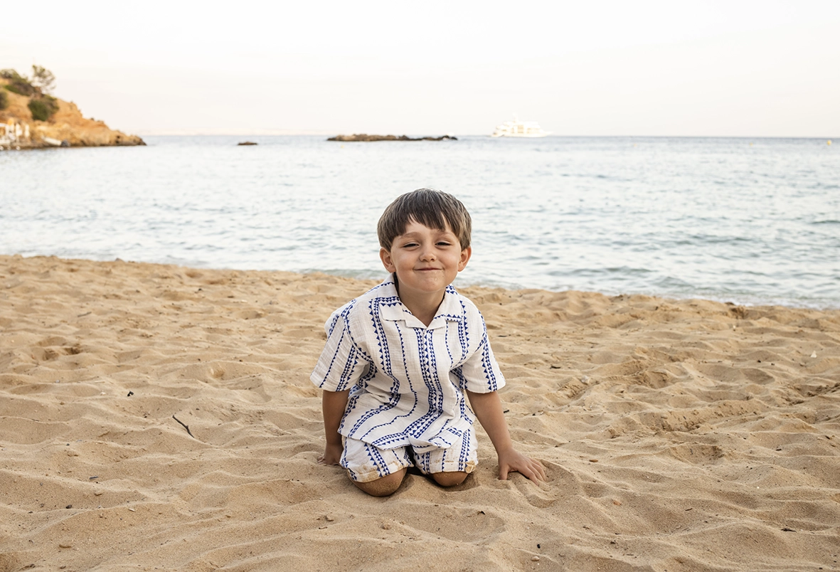 Fotógrafo familiar en Mallorca, niño pequeño sentado en la arena sonriendo frente al mar. Foto tomada por Inma del Valle.