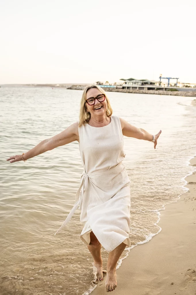 Fotógrafo familiar en Mallorca, mujer mayor caminando feliz por la orilla del mar con los brazos extendidos. Imagen de Inma del Valle.