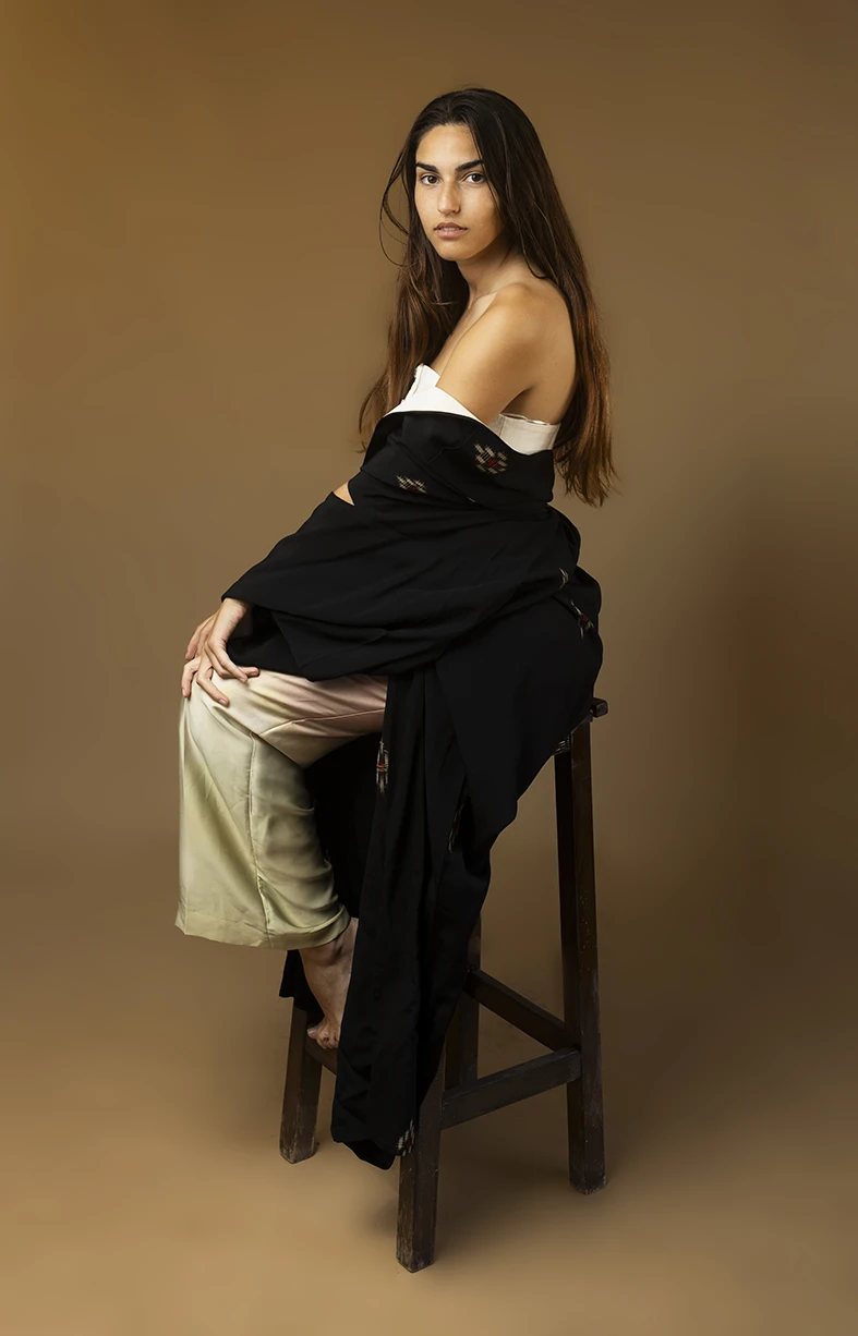 Model sitting in profile on a stool, wearing a creative layered outfit in a neutral studio setting. How you can become a model in Mallorca. Photo by Inma del Valle.