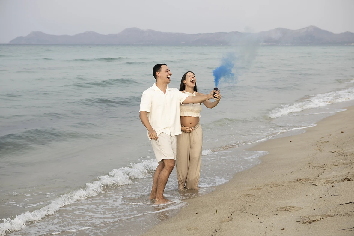 Pregnant couple celebrating on the beach in Mallorca with a blue smoke flare announcing they are expecting a boy.