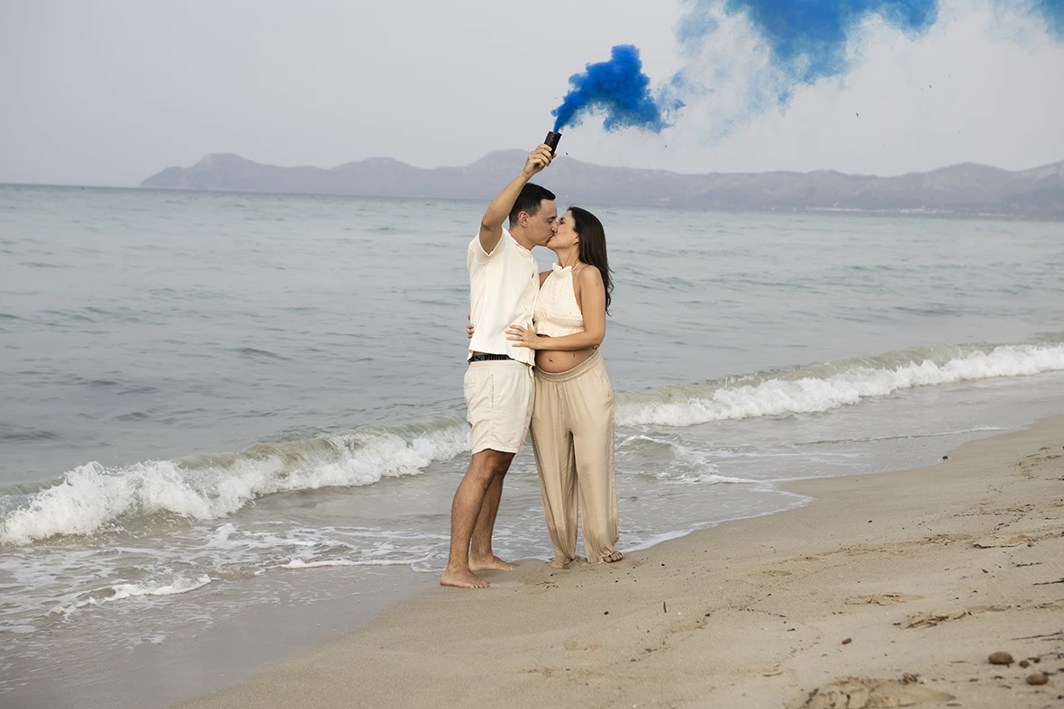 Pregnant couple embracing on the beach in Mallorca while raising a blue smoke flare.