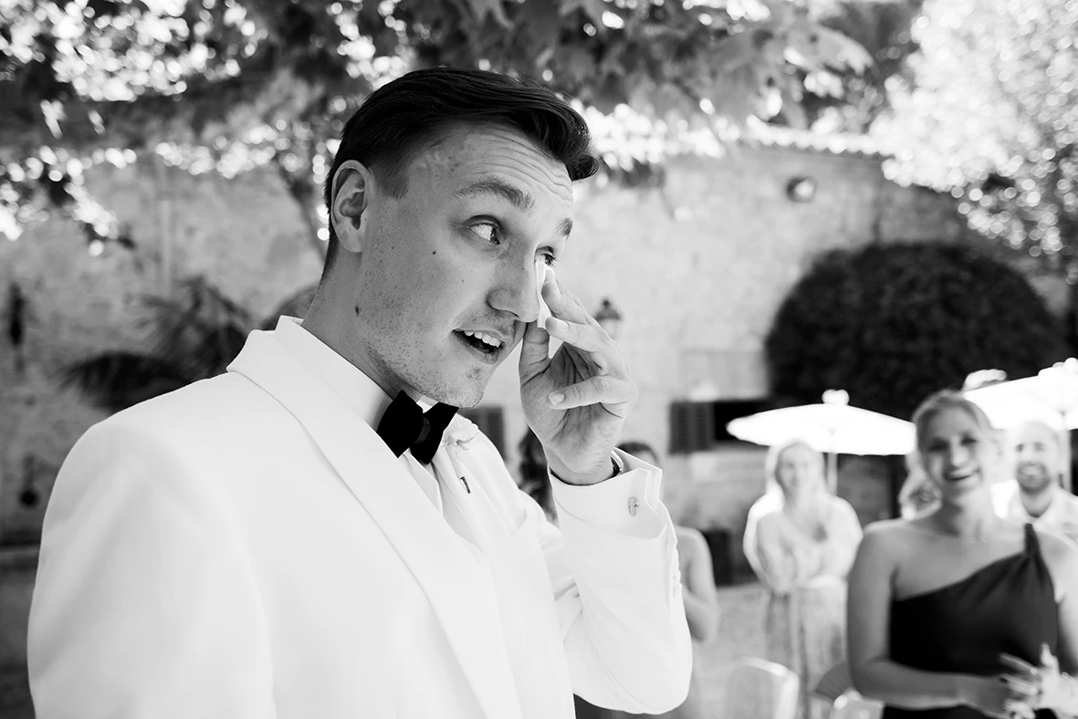 Groom in a white suit with black bow tie wiping a tear during his wedding in Mallorca.