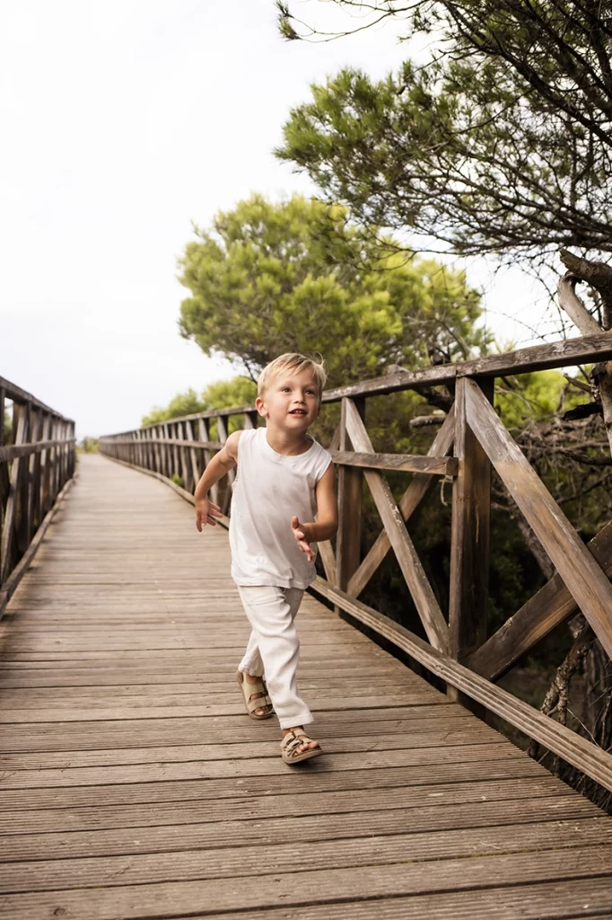 Sesión de fotos a familia en Mallorca realizada por Inma del Valle, niño corriendo con alegría por una pasarela de madera rodeada de árboles.