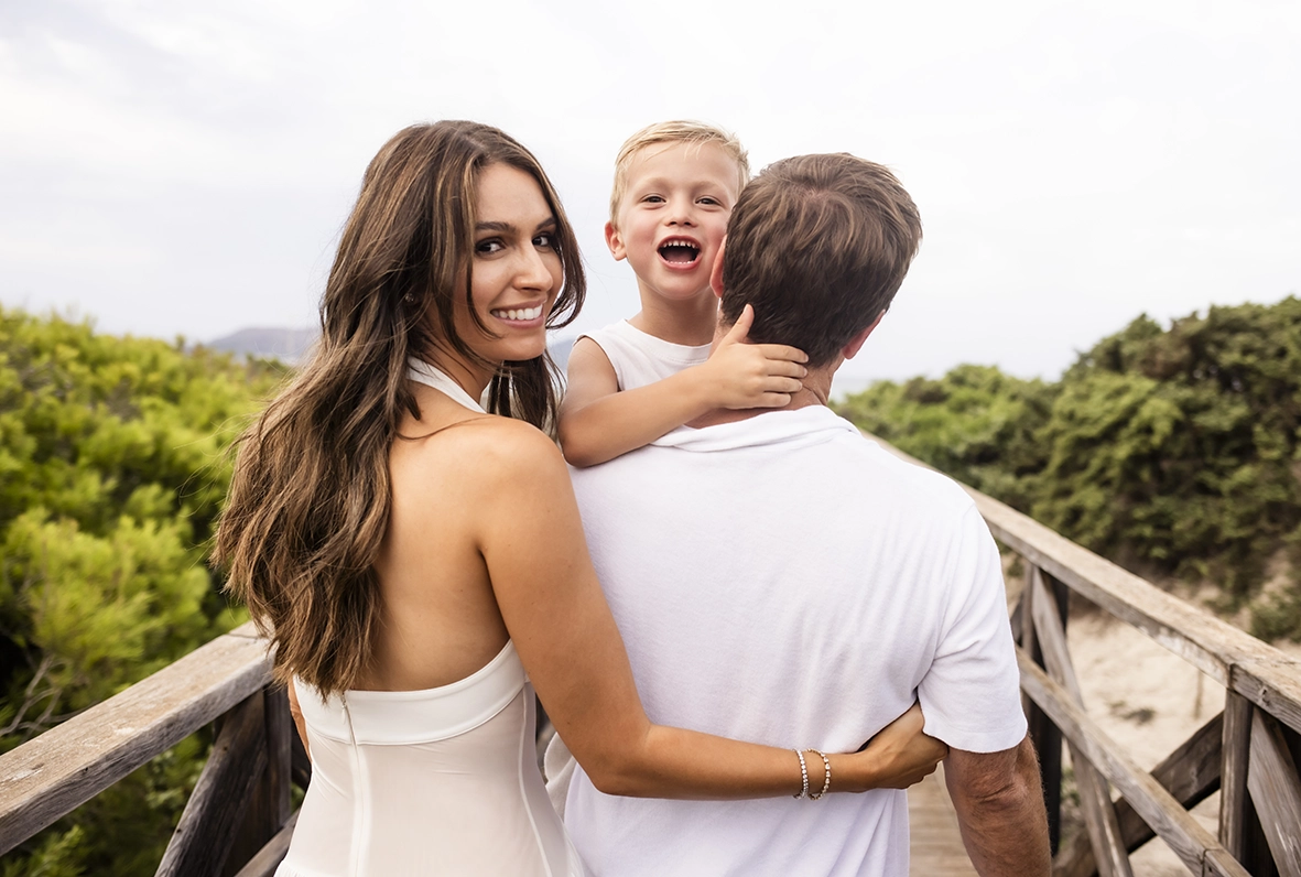 Sesión de fotos a familia en Mallorca realizada por Inma del Valle, madre mirando a cámara mientras abraza a su familia en una pasarela de madera.