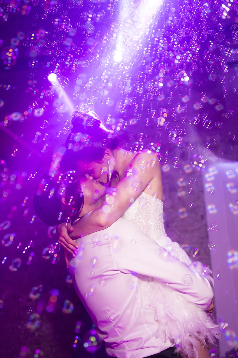 Novios besándose entre luces y burbujas, ella con su vestido de novia para la fiesta. Fotografía de Inma del Valle.