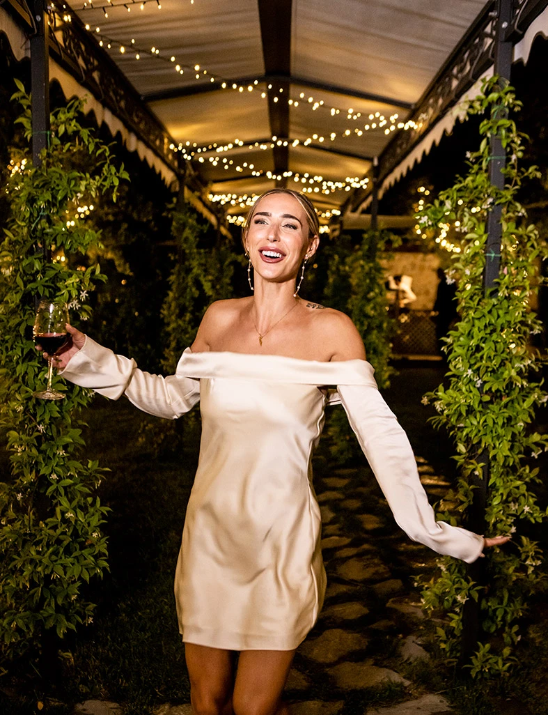 Novia caminando entre luces y vegetación con una copa en la mano y su vestido de novia para la fiesta. Fotografía de Inma del Valle.