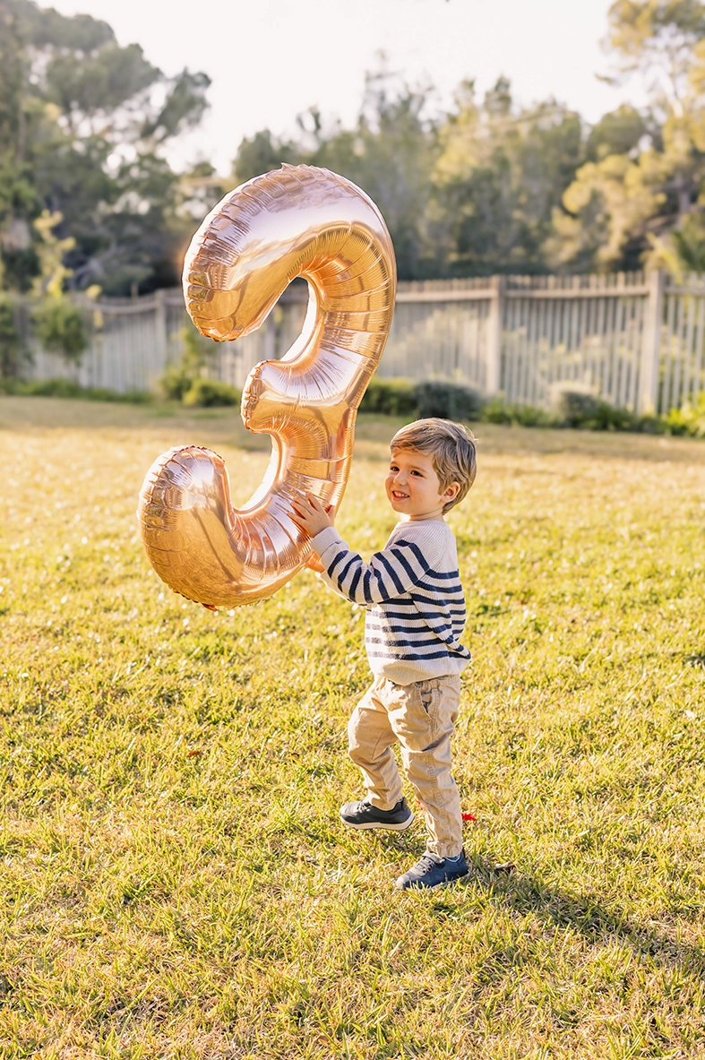 Niño jugando con un globo dorado en forma de número tres al aire libre, capturado por un fotógrafo para familias en Mallorca.