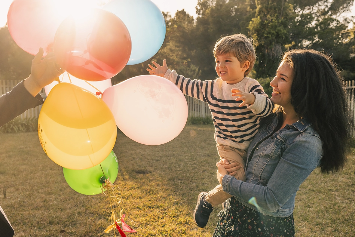 Niño en brazos de su madre alcanzando globos de colores al aire libre, capturado por un fotógrafo para familias en Mallorca.