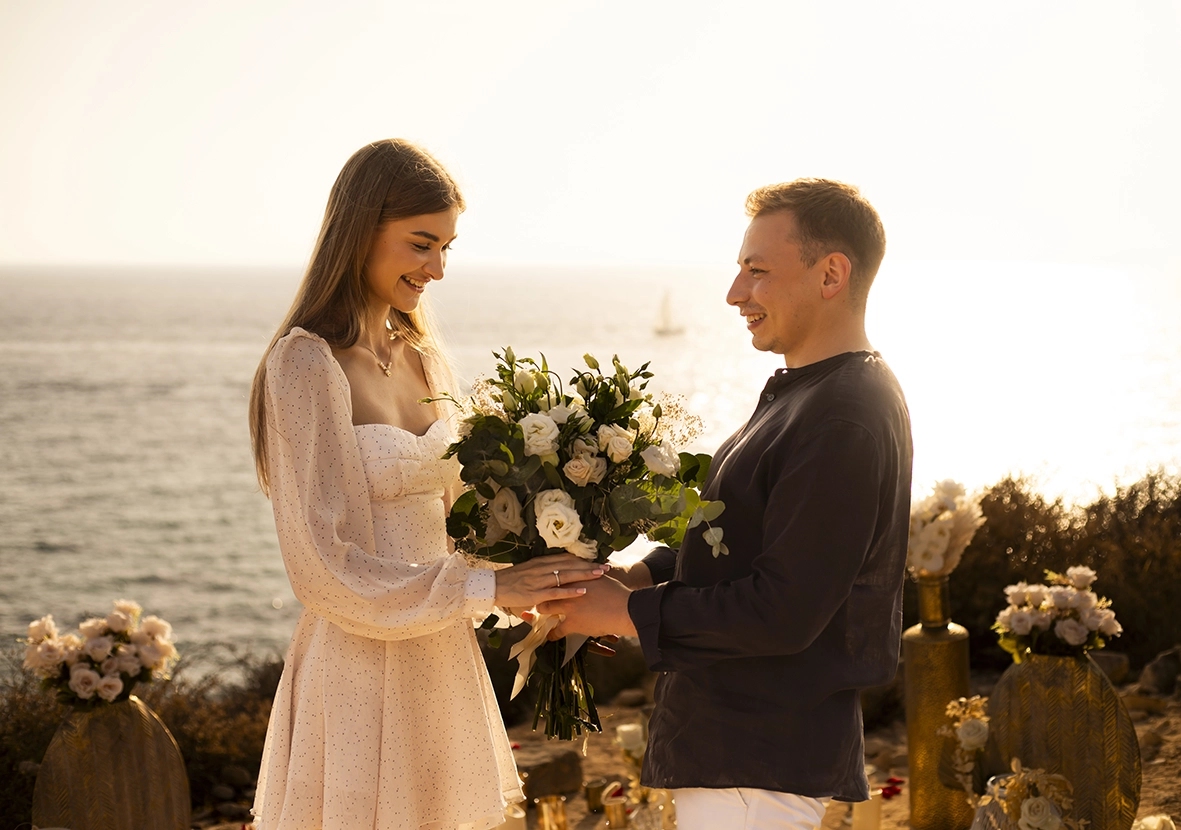 Couple smiling and holding a bouquet of flowers during the proposal by the sea, captured by a photographer for proposals in Mallorca.