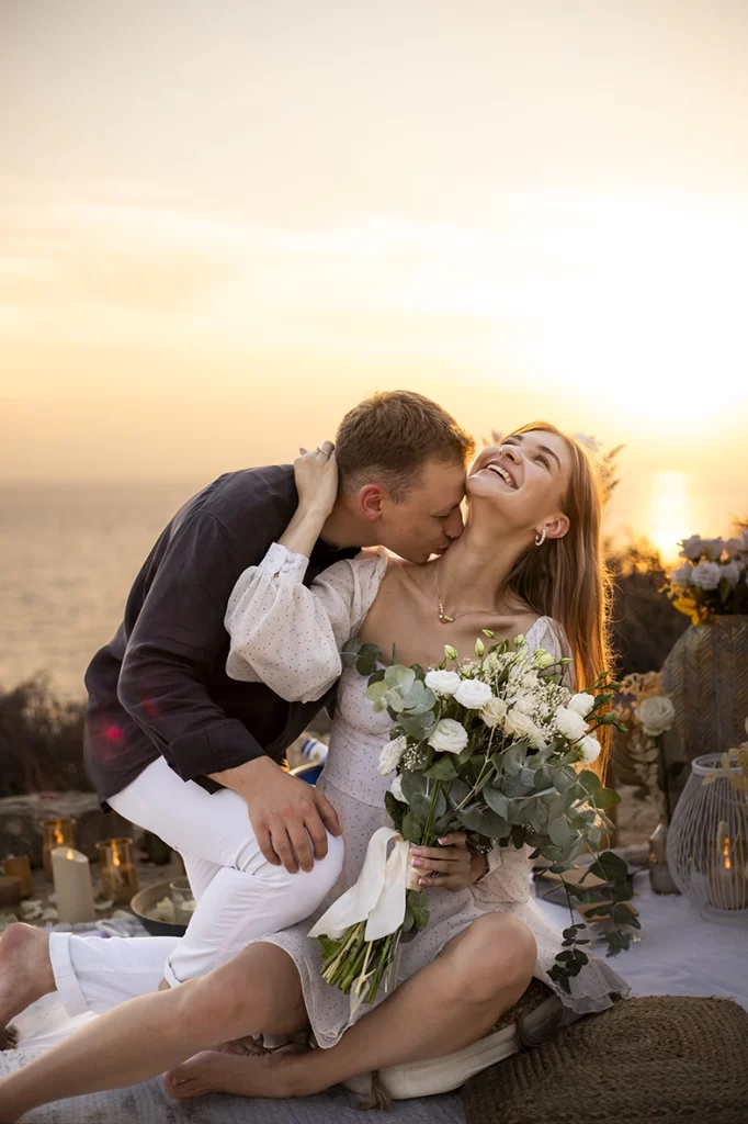 Happy couple during their proposal in Mallorca, he kisses his partner while she smiles holding a bouquet of flowers at sunset. Captured by a photographer for proposals in Mallorca.