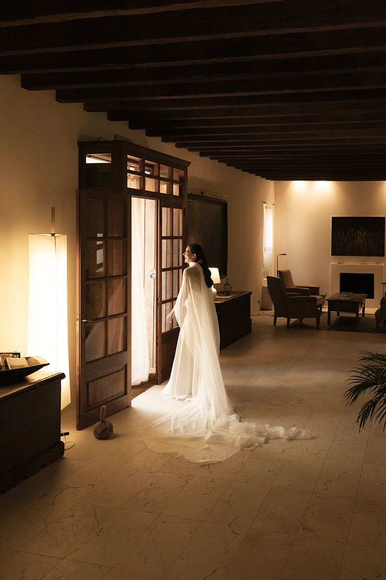 Bride in a white dress lit by warm indoor light, looking out through a wooden door – Wedding photographer in Mallorca – Inma del Valle -