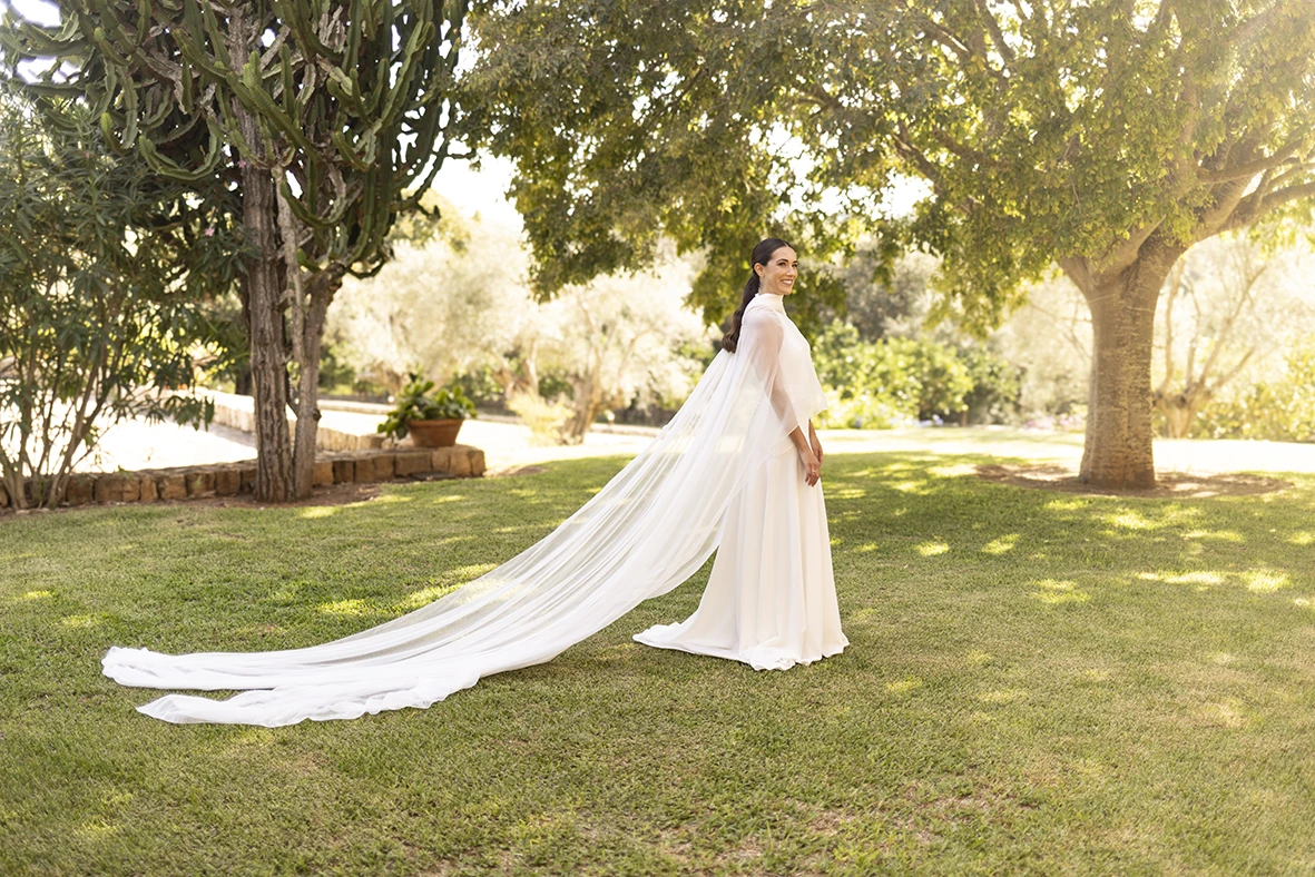Smiling bride in a white dress with a long veil posing on the grass under trees at sunset – Wedding photographer in Mallorca – Inma del Valle -