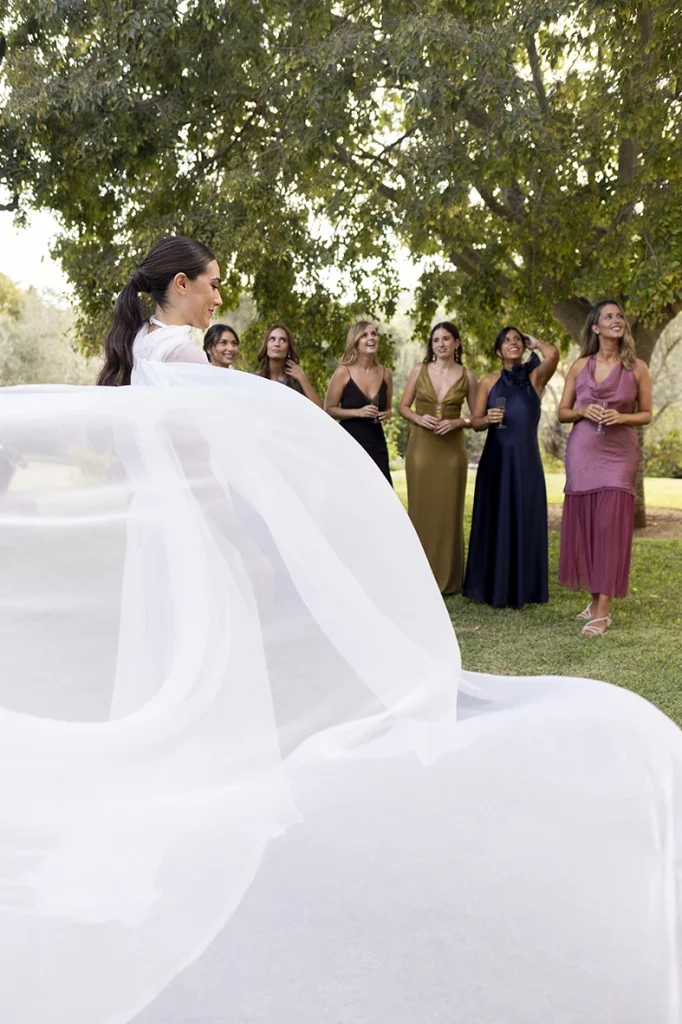 The bride in profile with her veil flowing in the wind, surrounded by smiling bridesmaids under a large tree – Wedding photographer in Mallorca – Inma del Valle -