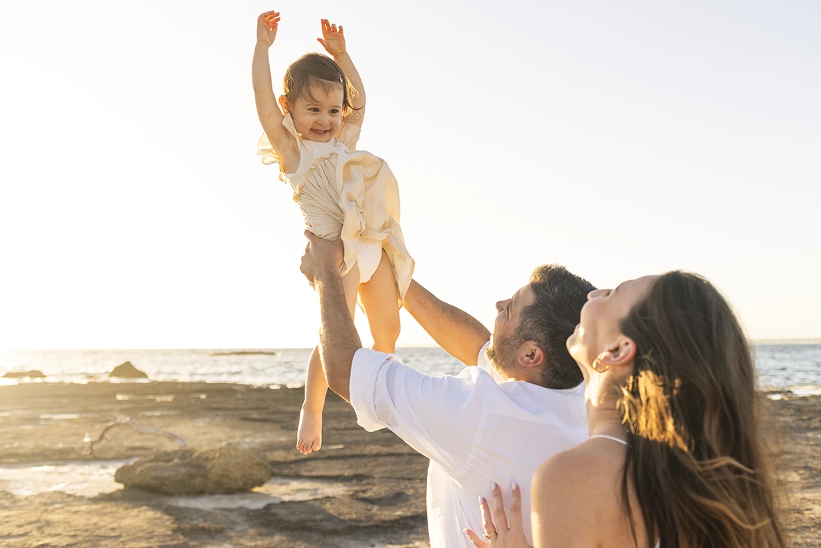 Padre levantando a su hija al cielo mientras la madre sonríe en una playa de Mallorca al atardecer