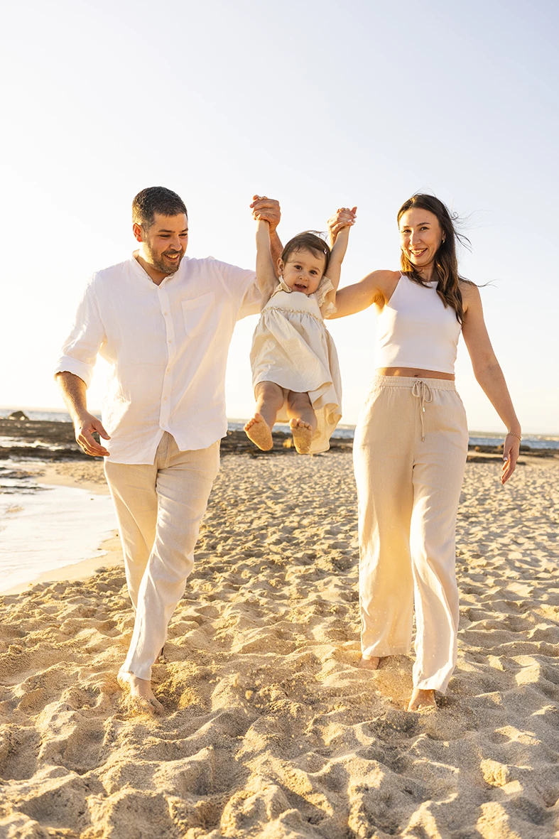 Padres caminando por la playa mientras balancean a su hija en una sesión de fotografía de familias en Mallorca