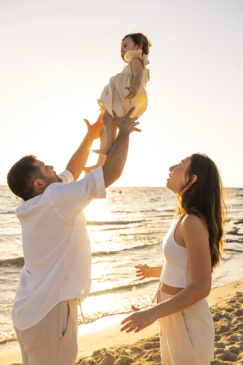 Padre levantando a su hija al cielo mientras la madre sonríe en la orilla del mar con luz dorada al atardecer