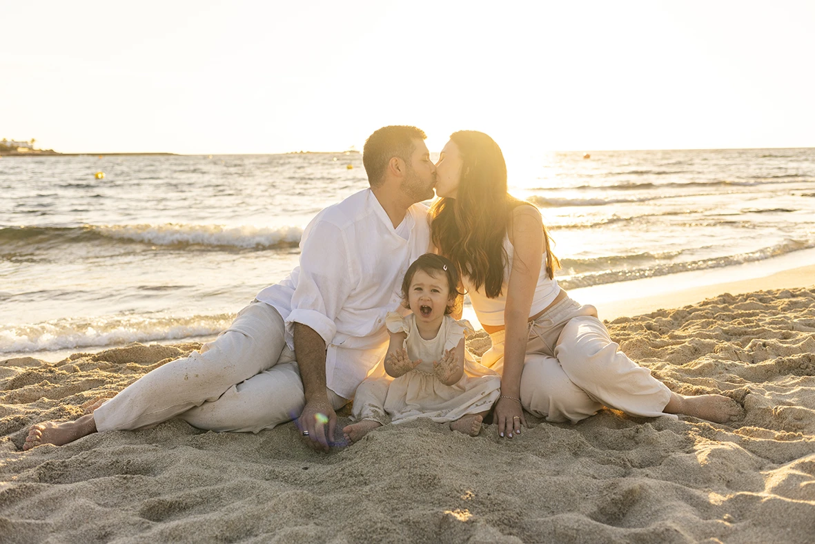 Padres besándose mientras su hija sonríe sentada entre ellos durante una sesión de fotografía de familias en Mallorca