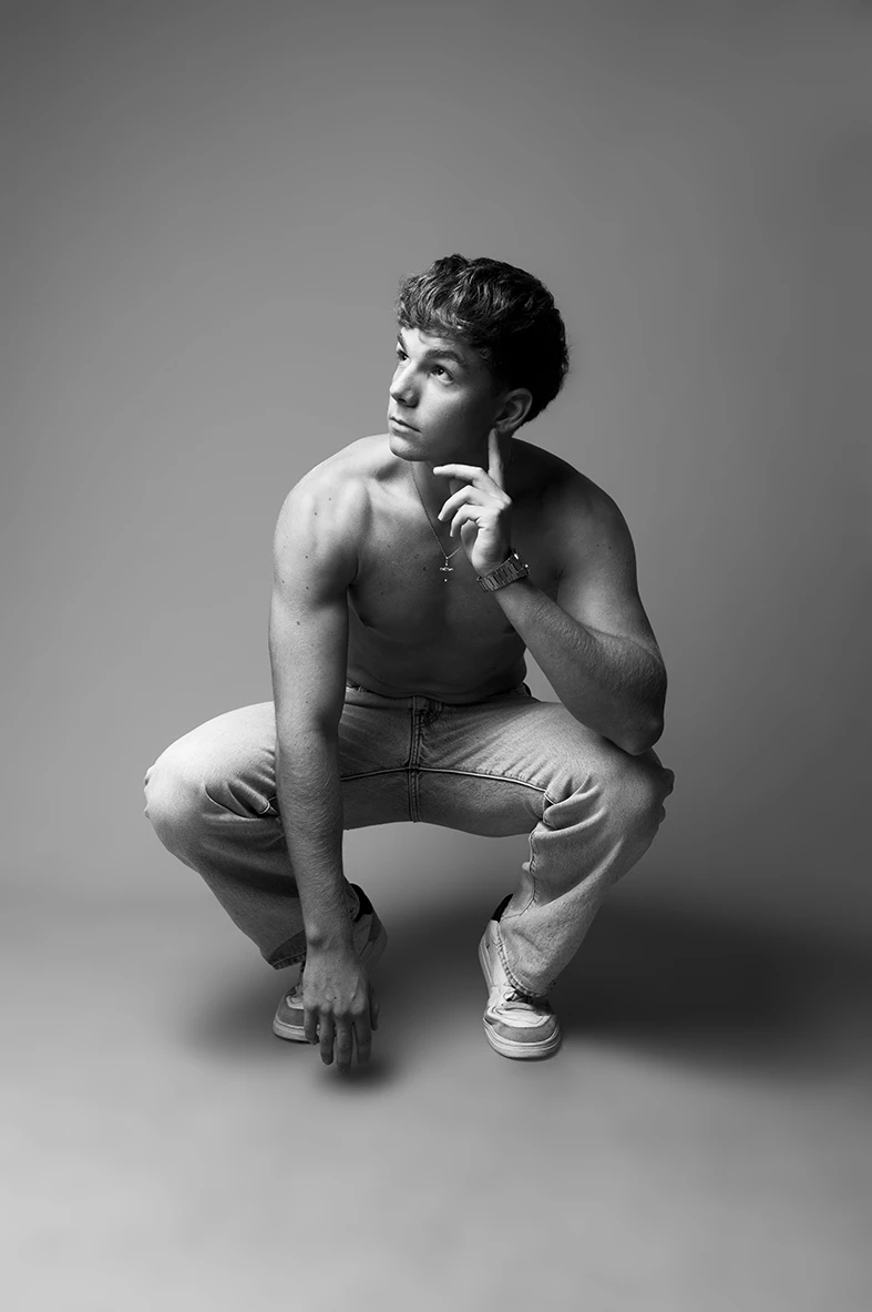 Modelo masculino agachado con expresión pensativa en foto de estudio en blanco y negro.