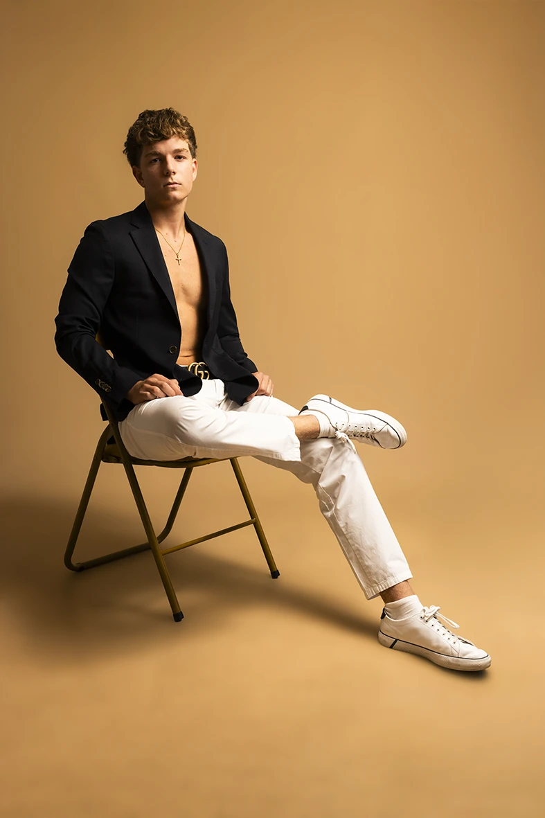 Male model wearing a black blazer posing on a chair during a studio photoshoot.