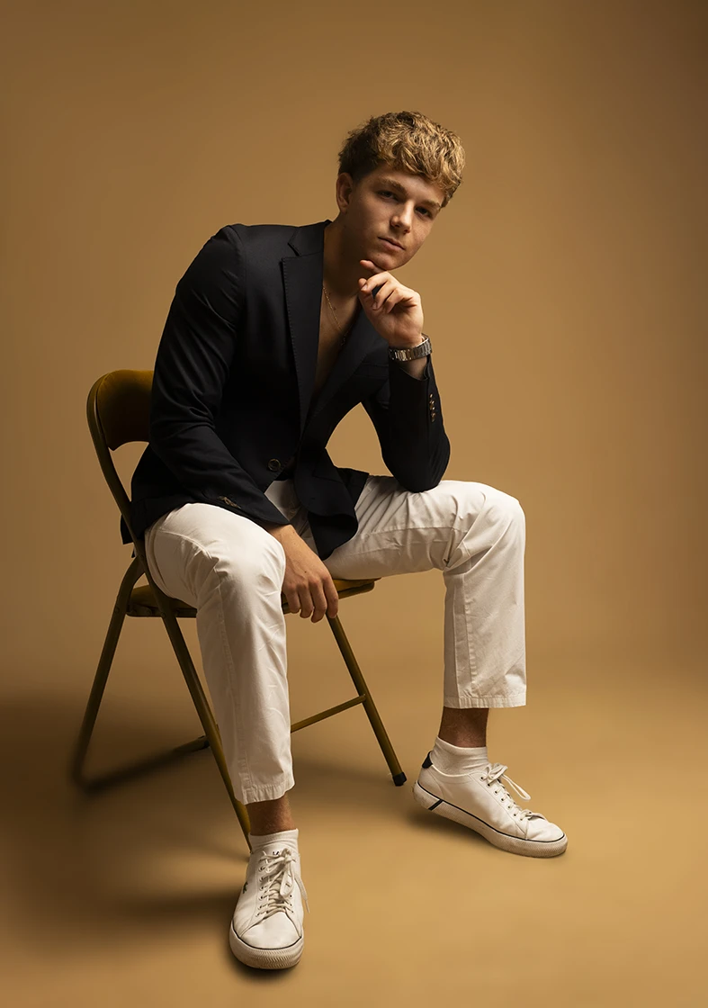 Male model seated with black blazer and serious expression in beige studio setting.