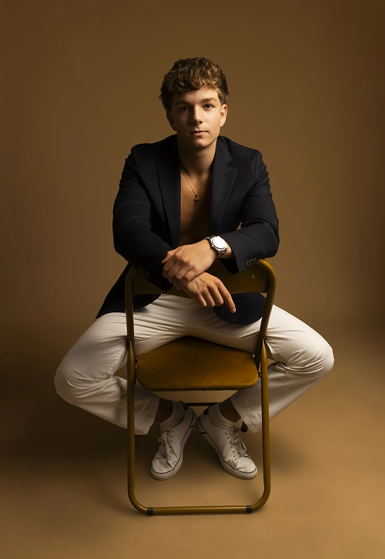 Male model sitting backwards on a chair wearing a black blazer and white pants.