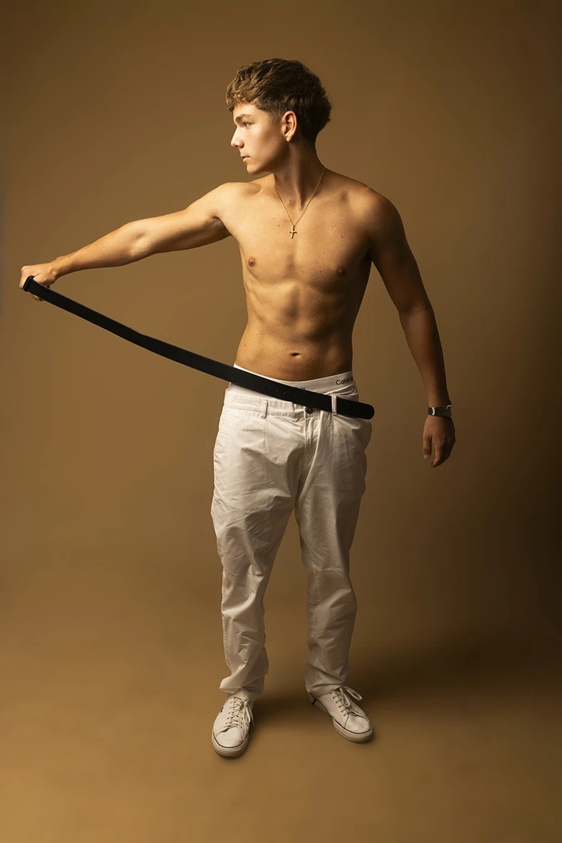 Shirtless male model playing with belt in studio with beige background.