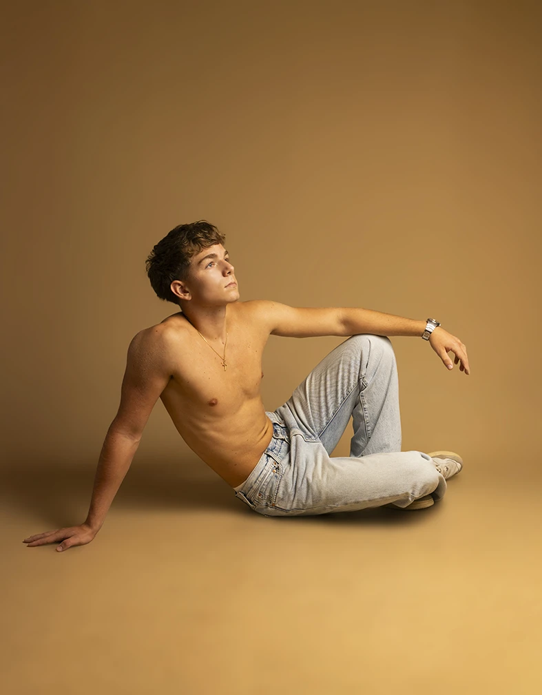 Shirtless male model posing on the floor in light jeans during studio photoshoot.