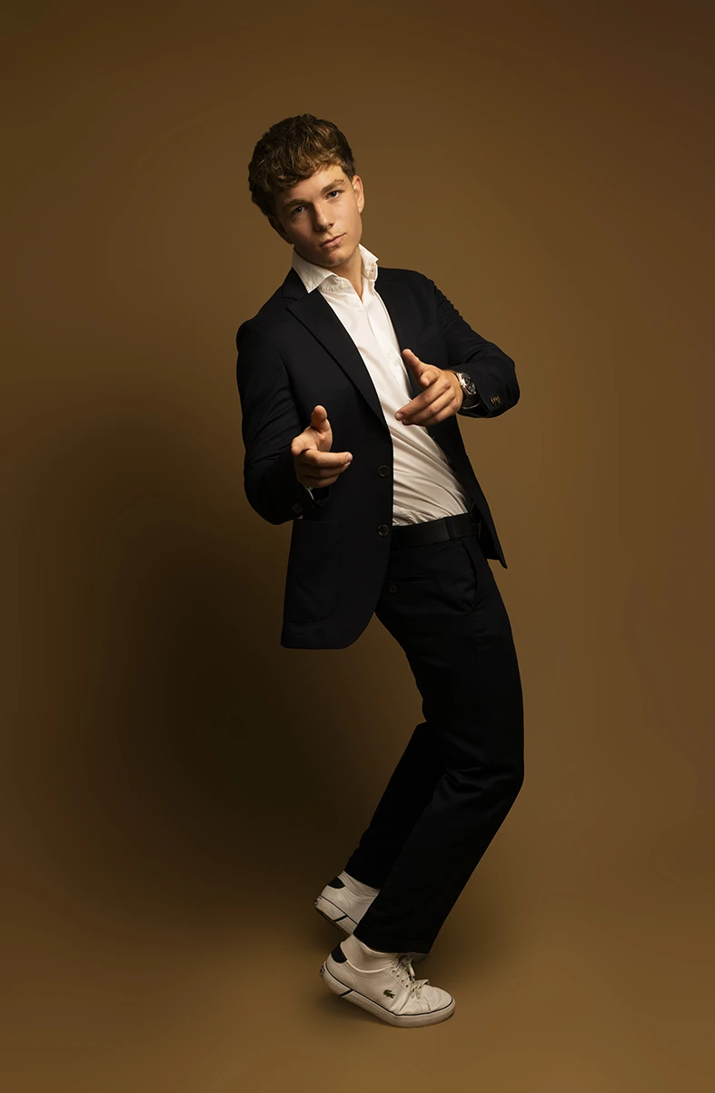 Male model in black suit striking fun pose with finger guns in studio.