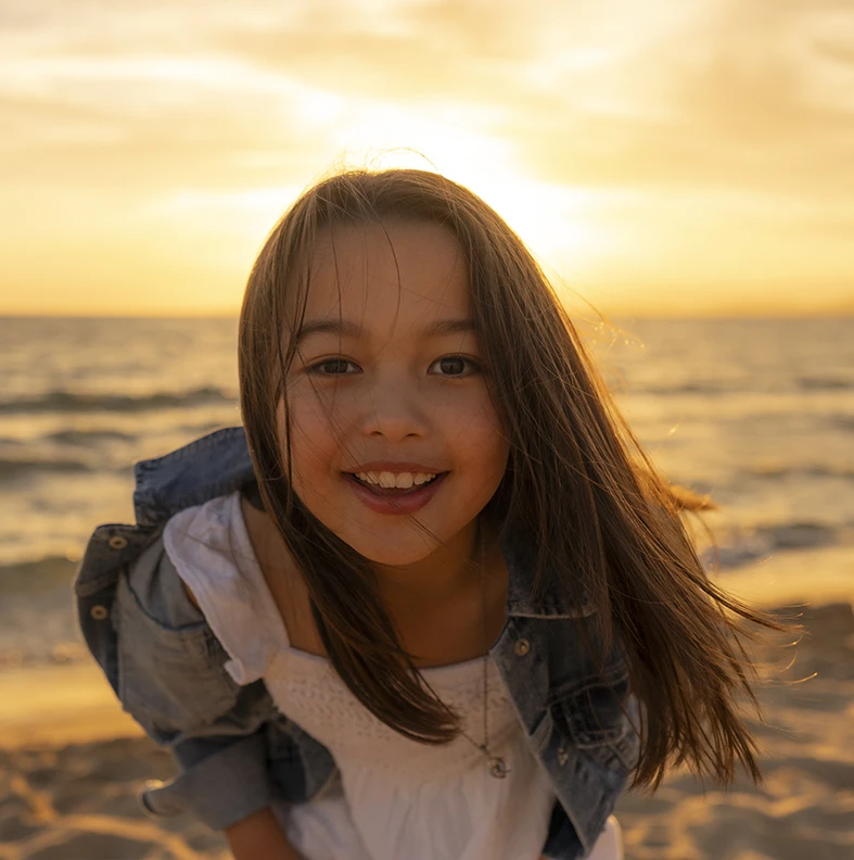 Primer plano de una niña sonriendo con el sol poniéndose detrás, en una playa de Mallorca. Fotografía realizada por Inma del Valle, fotógrafa en Mallorca.