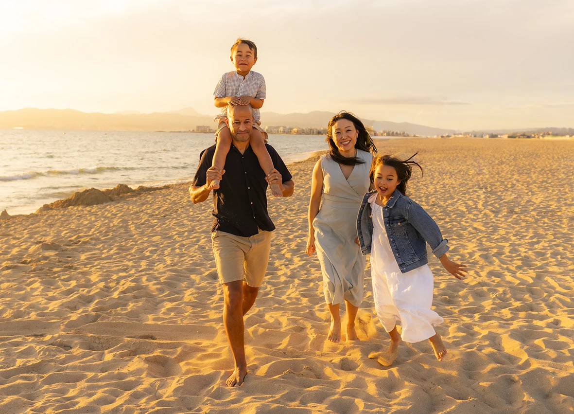Familia corriendo por la arena durante el atardecer en la playa de Mallorca. El padre lleva al niño sobre los hombros. Foto realizada por Inma del Valle, fotógrafa en Mallorca.